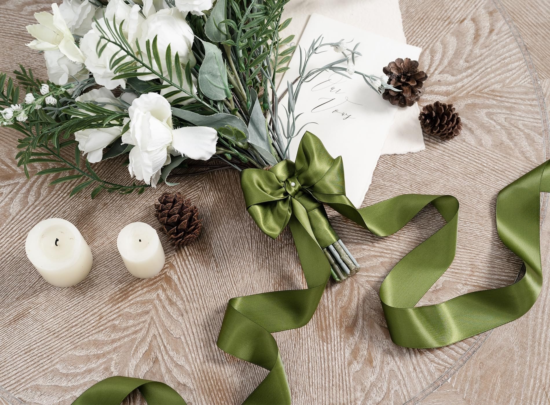 Green satin ribbon tied around a bouquet of white flowers with pinecones and candles on a wooden table