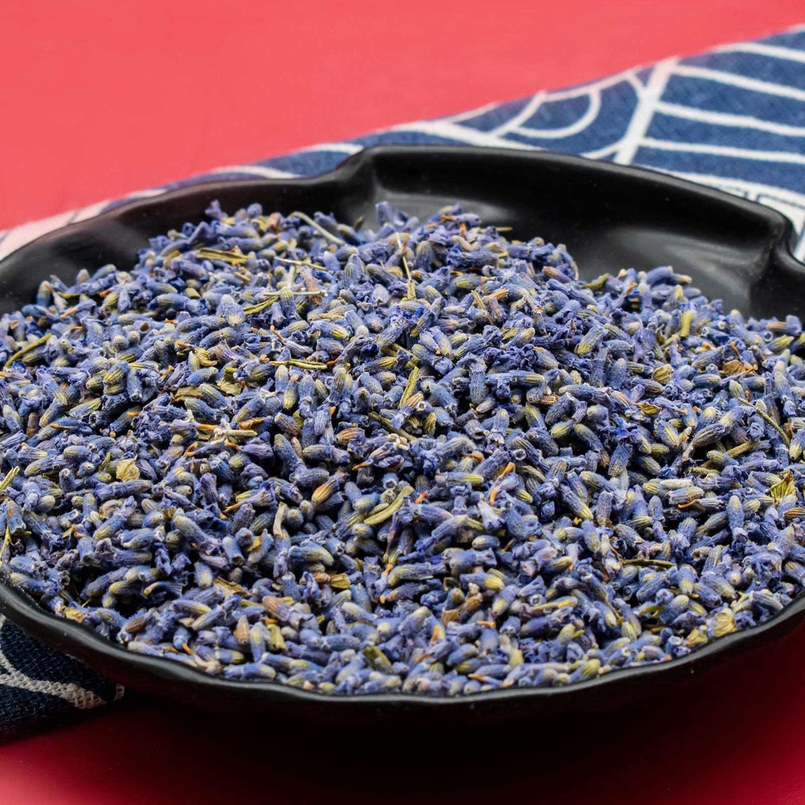 Close-up of dried lavender buds in a black bowl on a red background with a blue and white striped cloth.