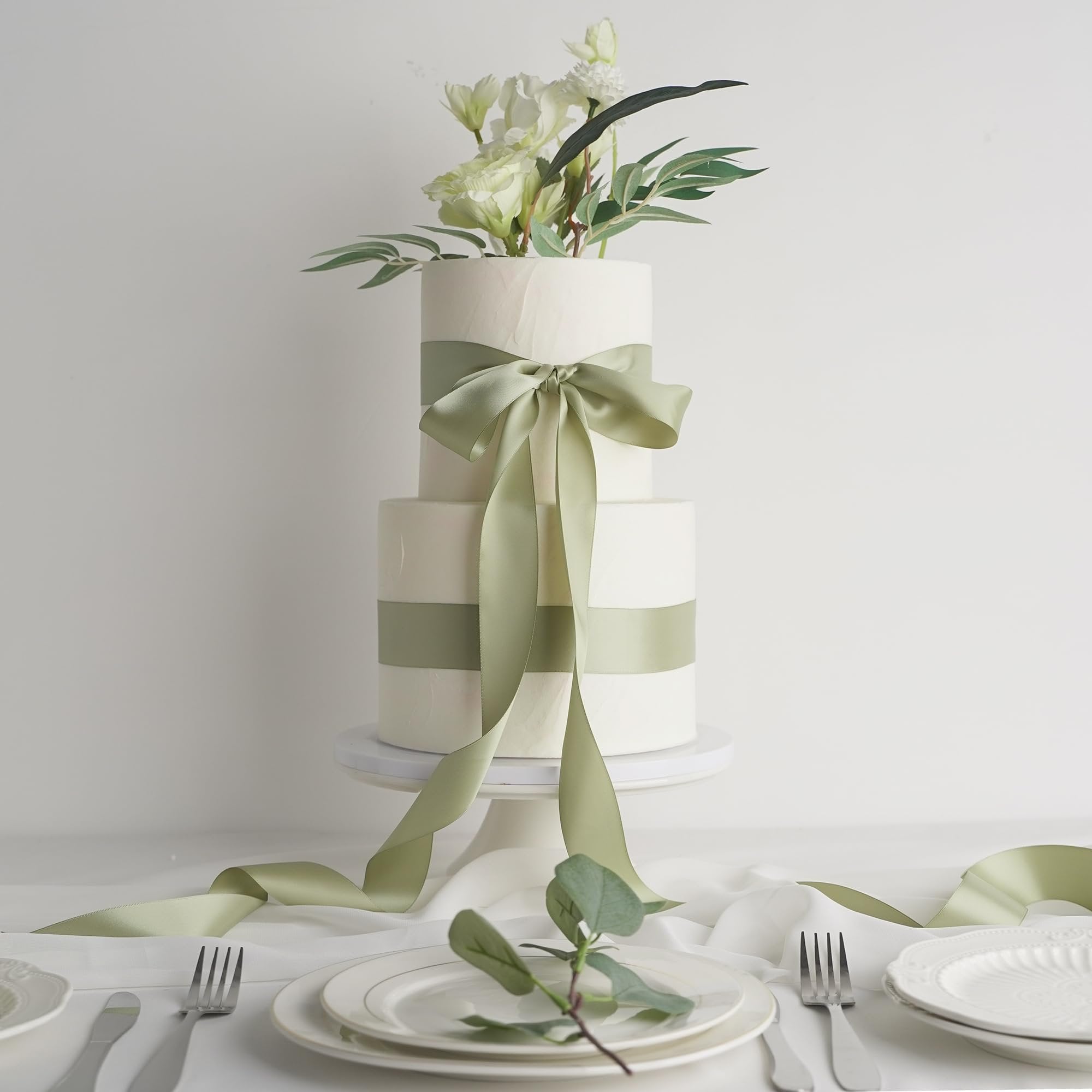 Two-tier white cake decorated with dusty sage satin ribbon and floral arrangement on a cake stand.