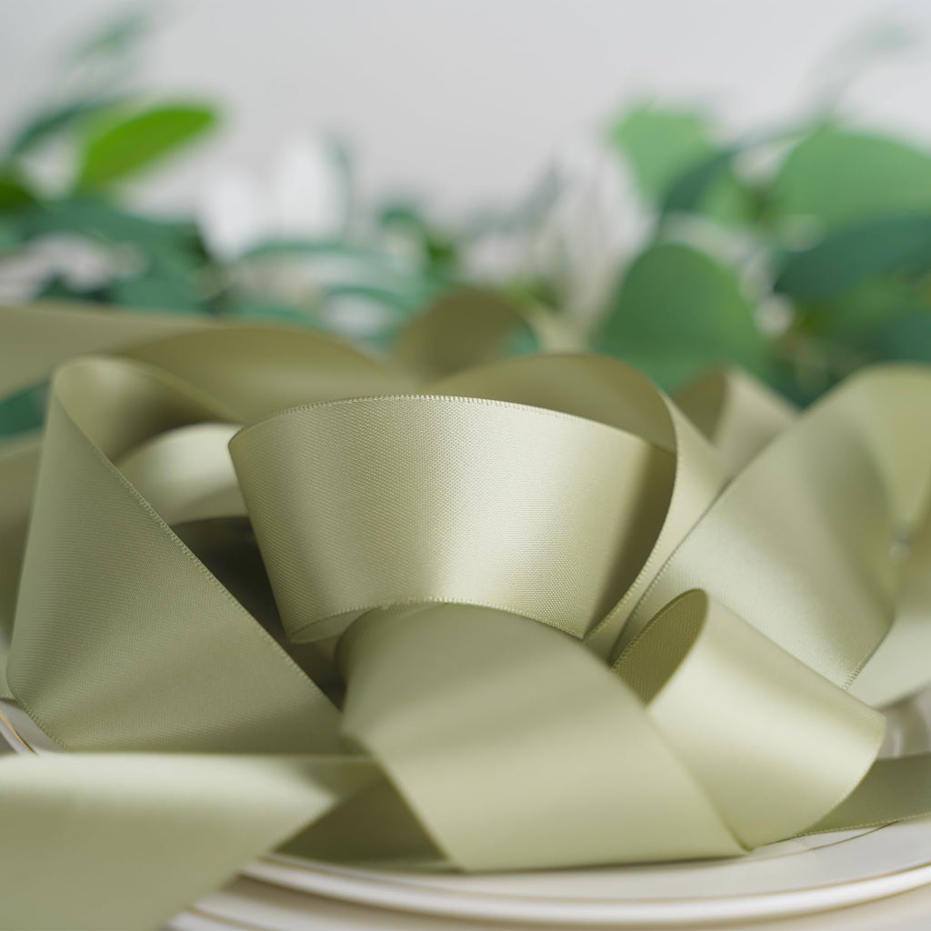 Dusty sage satin ribbon coiled on white plates with blurred green foliage in the background