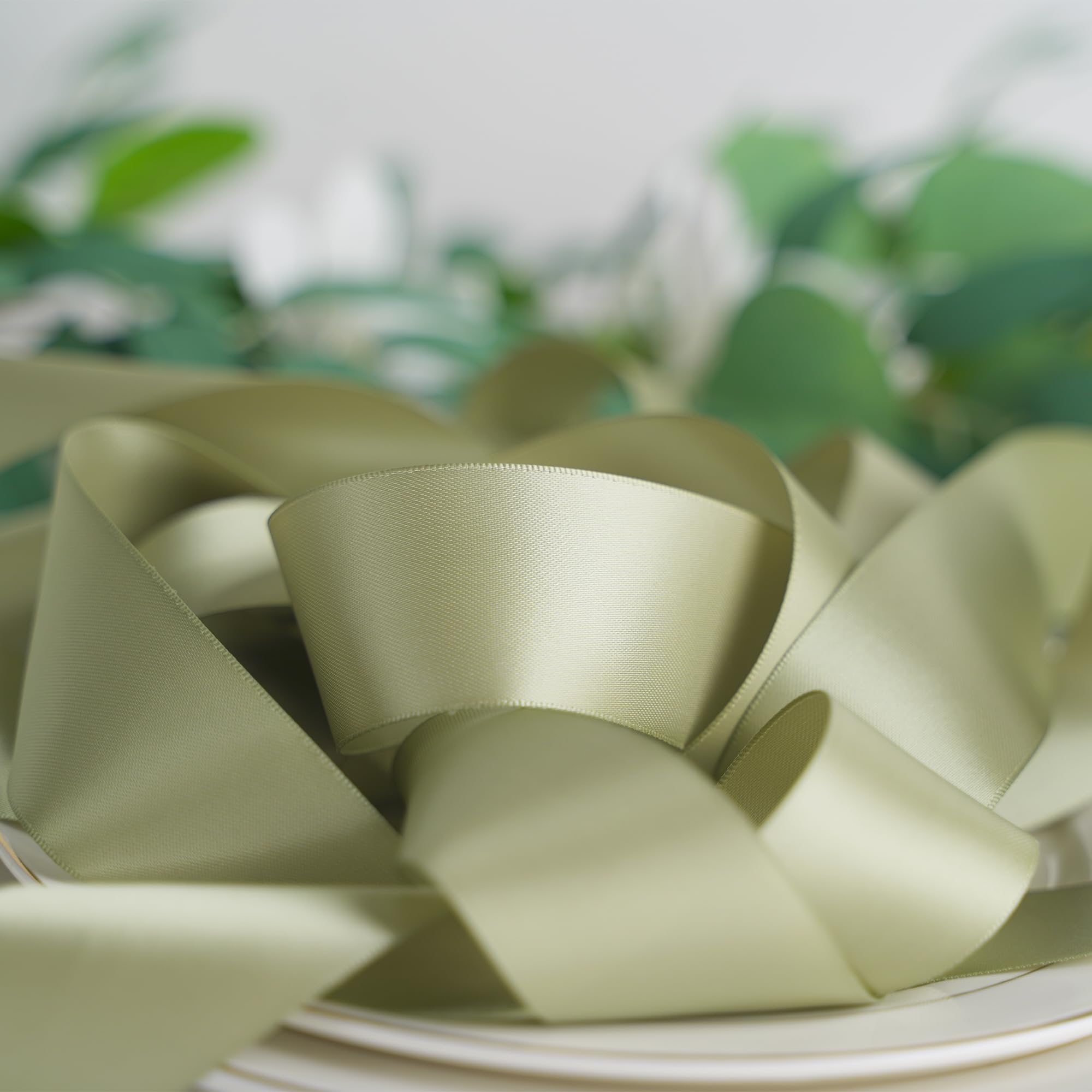 Dusty sage satin ribbon coiled on white plates with blurred green foliage in the background