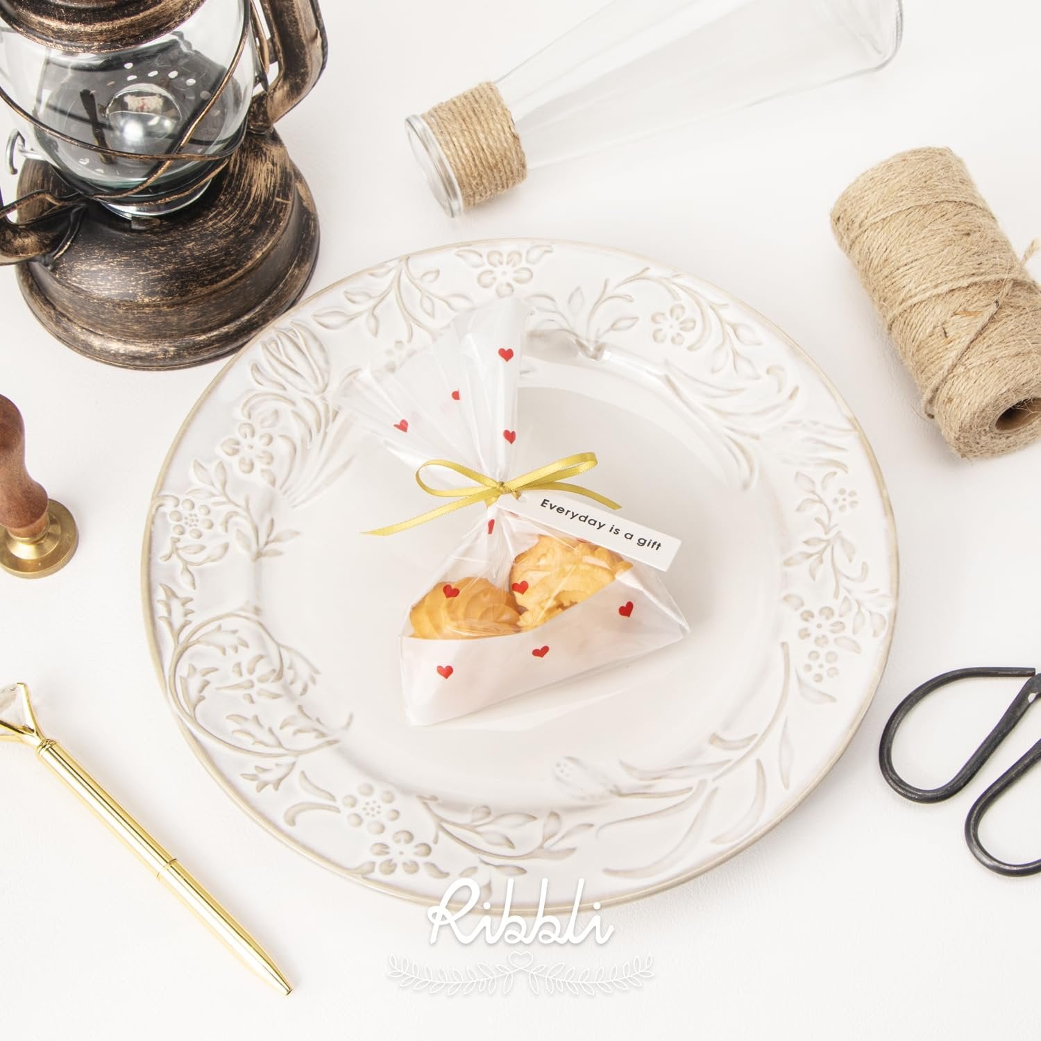 Gift bag with cookies on a decorative plate, surrounded by craft supplies and a vintage lantern.