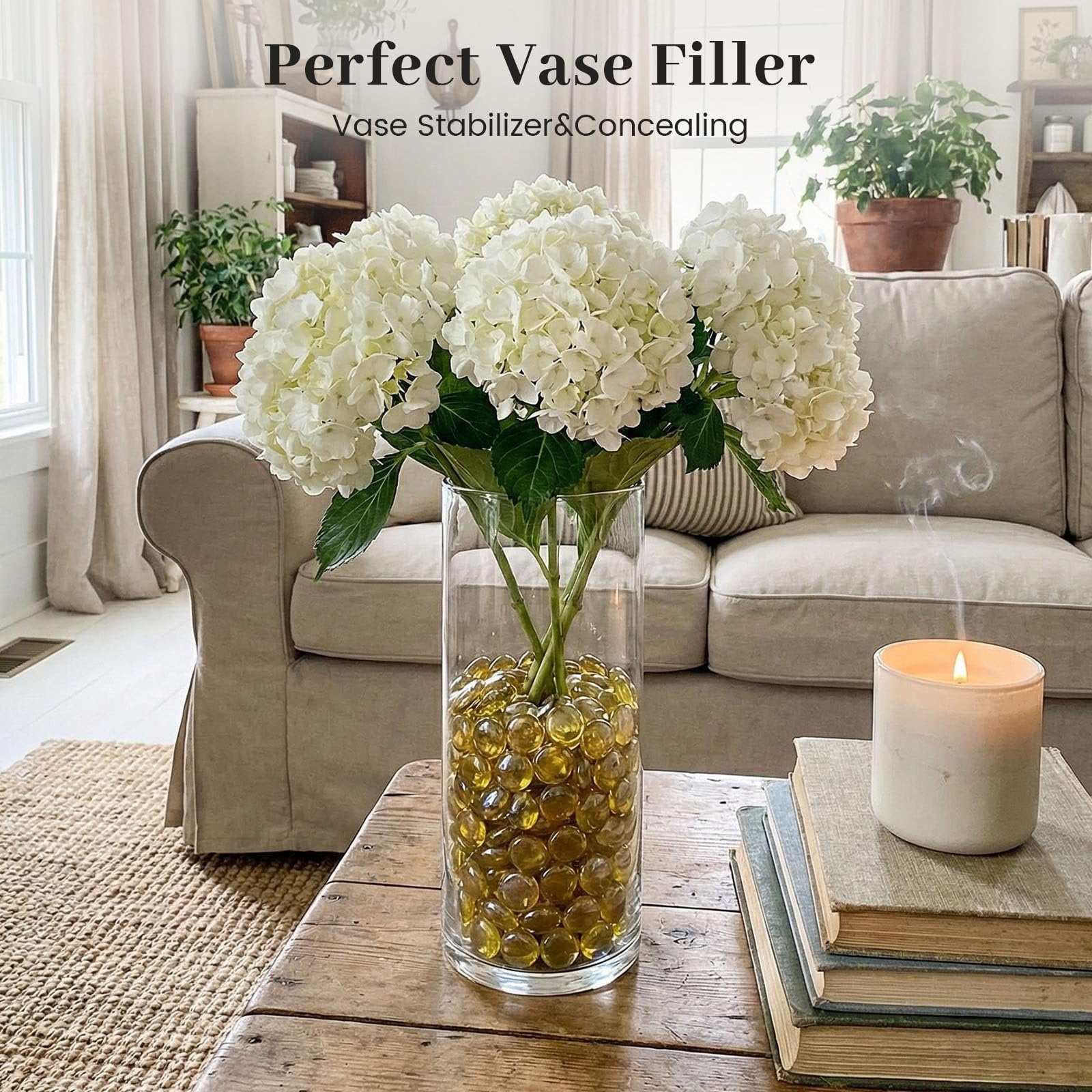 Amber glass marbles in a clear vase with white hydrangeas on a wooden table, next to books and a lit candle.