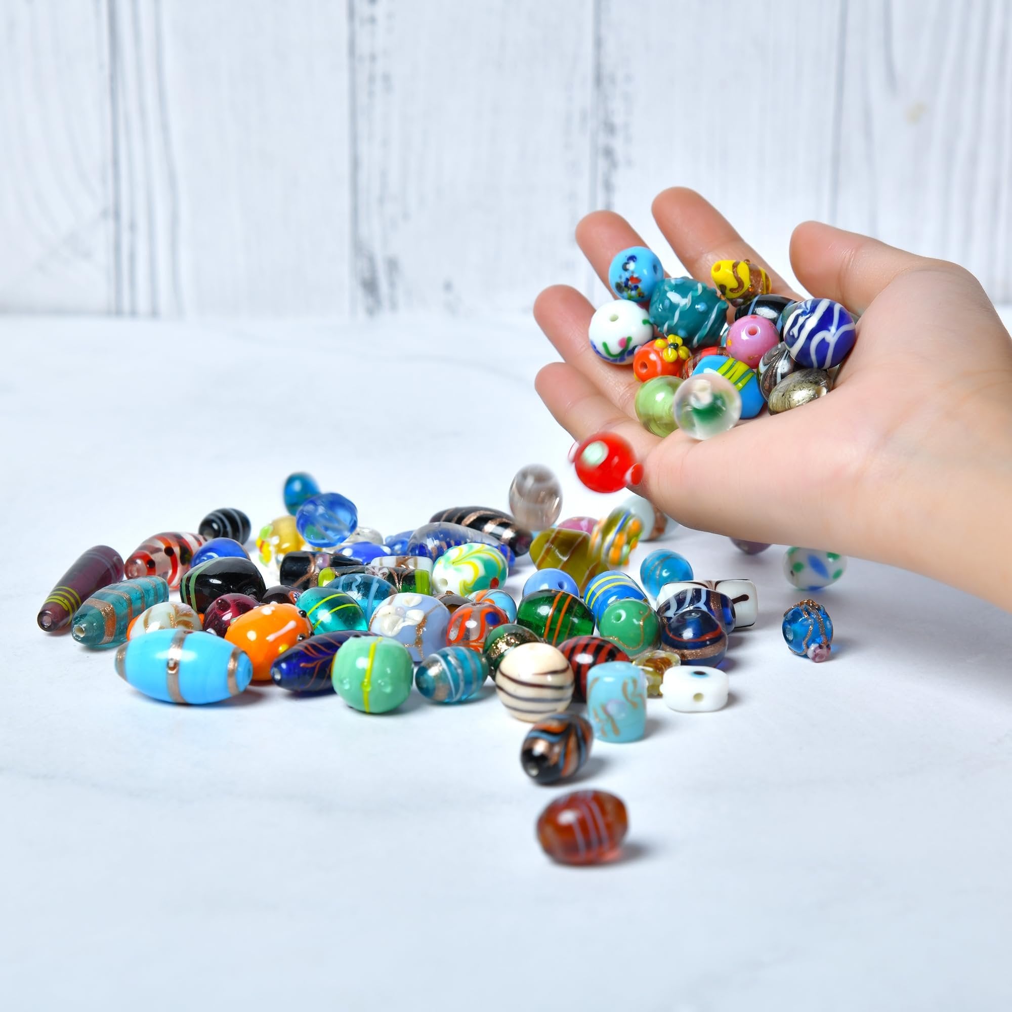 Hand holding assorted colorful glass beads, with more beads scattered on a white surface and a light wood background.
