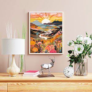 Framed paint by numbers artwork of a colorful mountain landscape with a river and flowers, displayed on a wall above a wooden dresser with decor.
