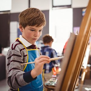 Boy wearing apron and painting on canvas at an easel in an art class
