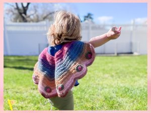 Toddler wearing a colorful butterfly-wing crochet shawl outdoors, facing away from camera.