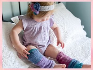 Toddler wearing knitted leg warmers and a headband with flowers, sitting on a bed.