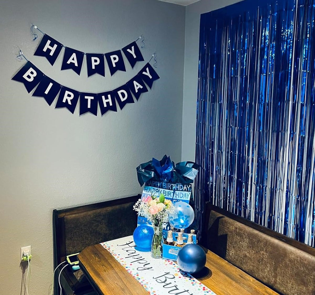 Party table setting with a navy blue 'Happy Birthday' banner hanging on the wall and blue metallic fringe curtains.