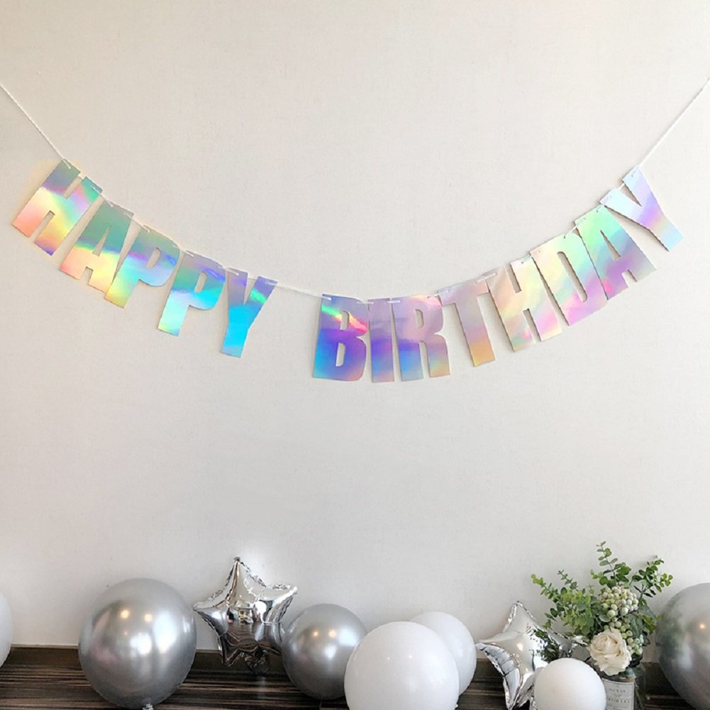 Holographic iridescent white 'Happy Birthday' banner strung on white cord, displayed against a white wall with silver and white balloons and floral arrangement below.