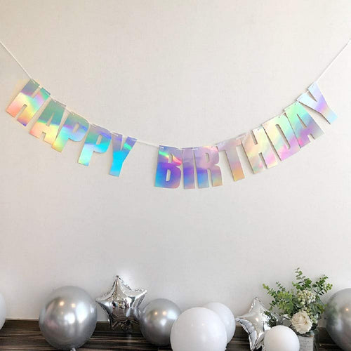 Holographic iridescent white 'Happy Birthday' banner strung on white cord, displayed above silver and white balloons and a floral arrangement.