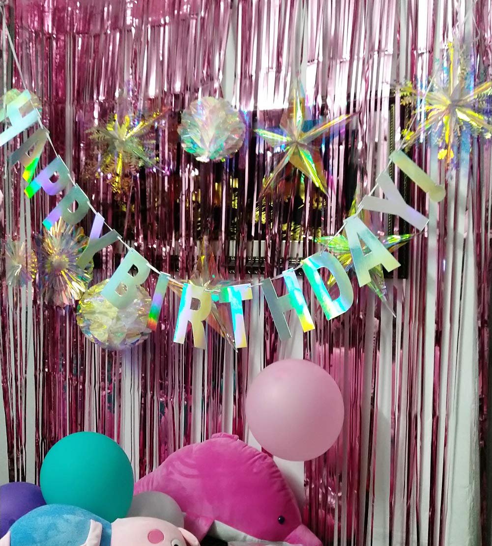 Holographic iridescent white 'Happy Birthday' banner hanging in front of a pink tinsel curtain, with balloons and plush toys below.