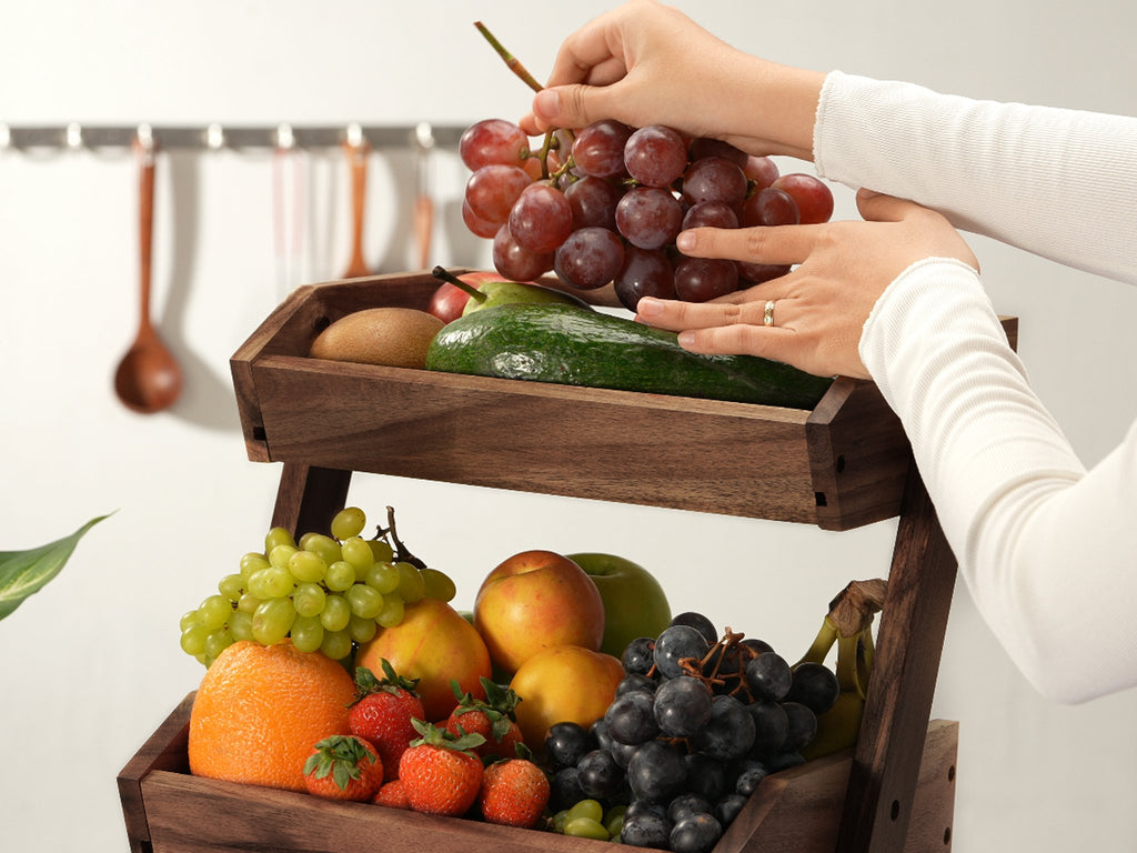 Two-tier wooden fruit basket filled with various fruits, with hands adding grapes to the top tier.