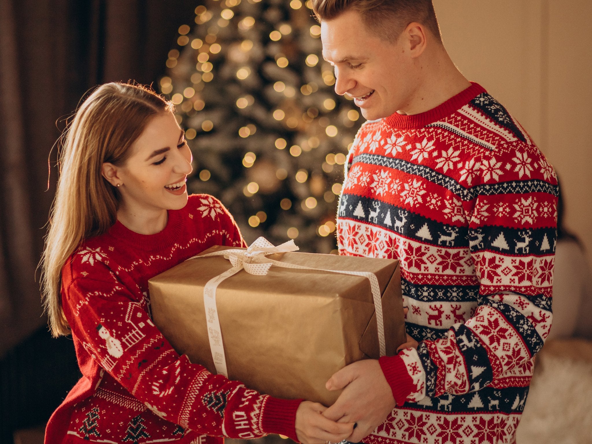 Couple in festive sweaters exchanging a wrapped gift in front of a decorated Christmas tree