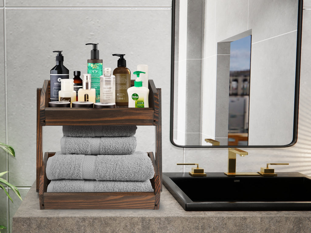 Two-tier dark wood bathroom organizer with toiletries on top shelf and folded gray towels on bottom shelf, next to a sink.