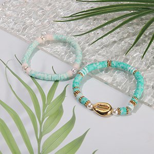 Two handmade bracelets with clay heishi beads, pearls, and gold accents on a light background with green leaves.
