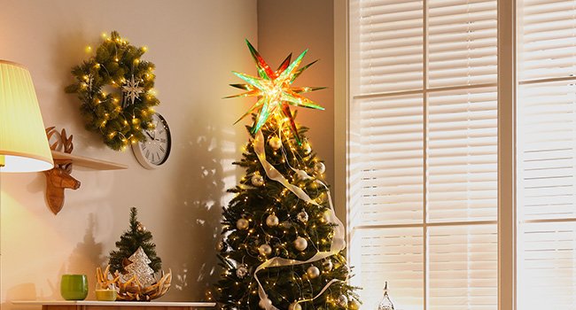 Christmas tree topped with a multi-pointed star, decorated with ornaments and lights, next to a wall with a wreath and clock.