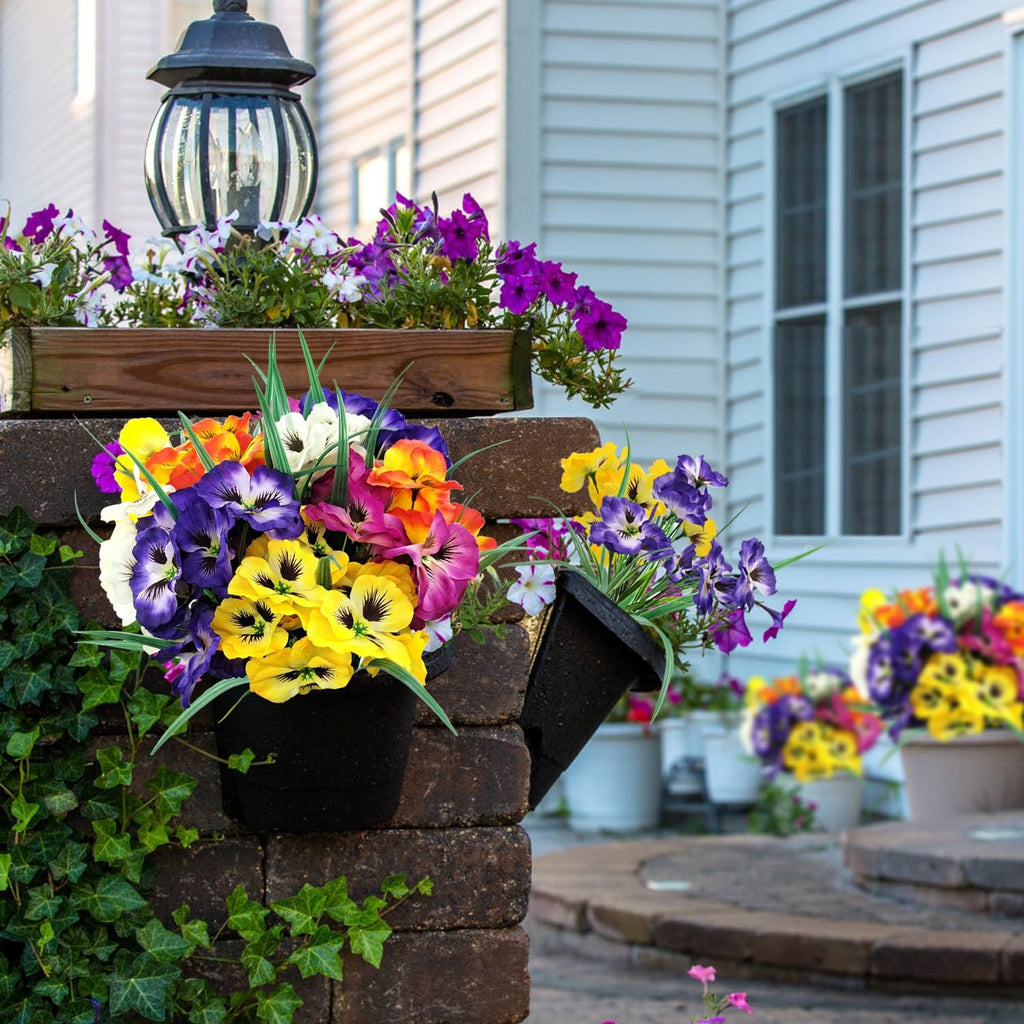 Artificial pansy flowers in multiple colors arranged in hanging planters and pots on a brick pillar and patio.