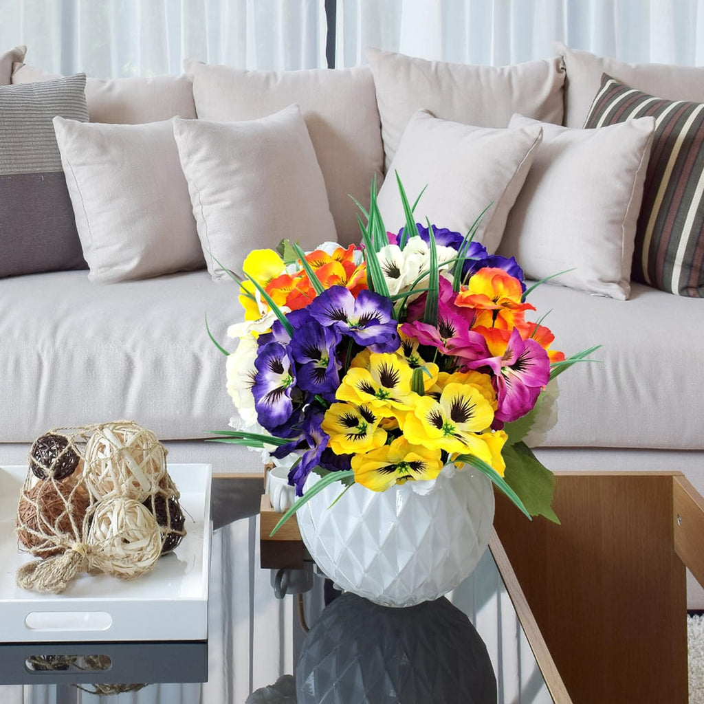 Colorful artificial pansy flowers in a white vase on a glass table, with a sofa in the background.