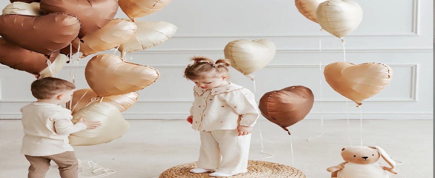 Two children and a teddy bear surrounded by heart-shaped balloons in sage green and beige.