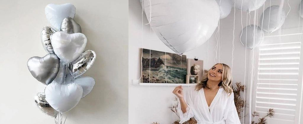A woman in a white robe looks up at a cluster of silver and white heart-shaped balloons floating indoors.