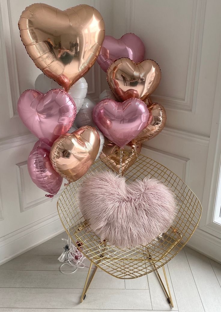 Group of heart-shaped foil balloons in rose gold, pink, and white, arranged around a gold wire chair with a pink heart-shaped fluffy pillow.