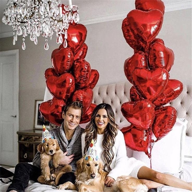 Couple with two golden retrievers and red heart balloons on a bed, wearing party hats