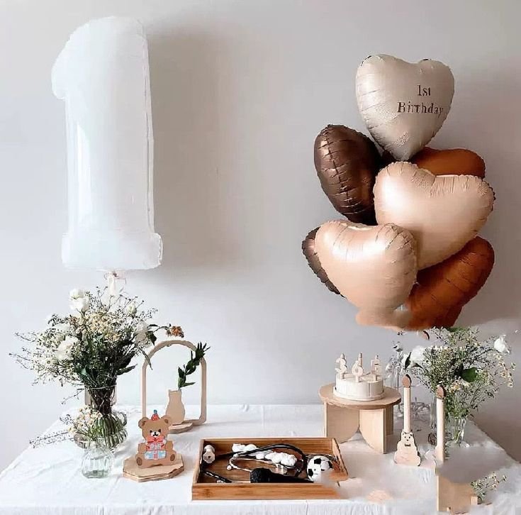 White number 1 balloon and heart-shaped balloons arranged on a table with a cake and decorations for a 1st birthday celebration.