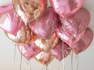 Cluster of rose gold and pink foil heart-shaped balloons with silver ribbons against a white background.
