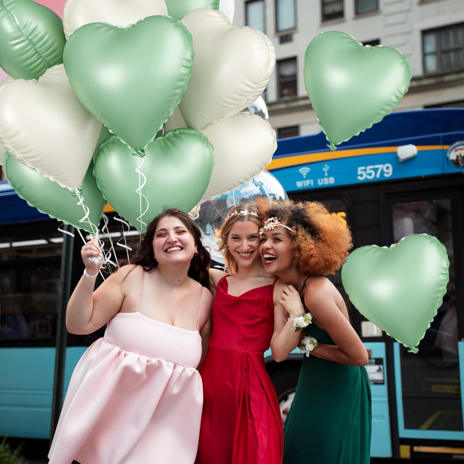 Three women in formal dresses holding sage green and cream heart-shaped balloons with a bus in the background.