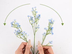 Hands arranging light blue artificial baby's breath flowers with green stems against a white background.