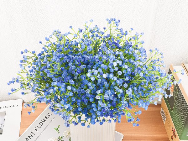 Blue artificial baby's breath bouquet with flexible stems in a white vase, placed on a wooden surface with books and magazines.
