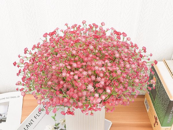 Pink artificial baby's breath bouquet in a white vase on a wooden surface, with books in the background.
