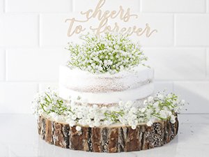 Two-tier naked cake decorated with artificial baby's breath and a wooden 'cheers to forever' topper, on a wood slice stand.