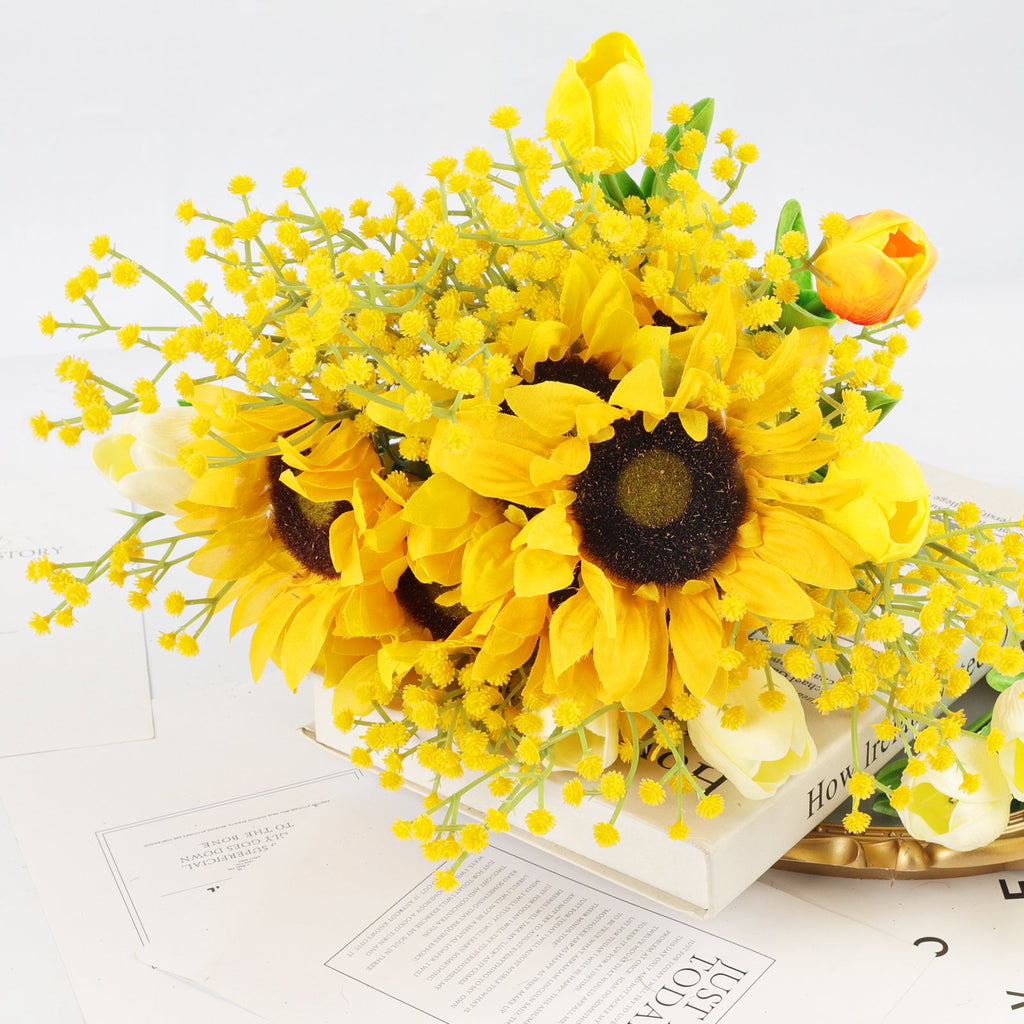 Artificial sunflower and tulip bouquet with yellow filler flowers arranged on books and papers.