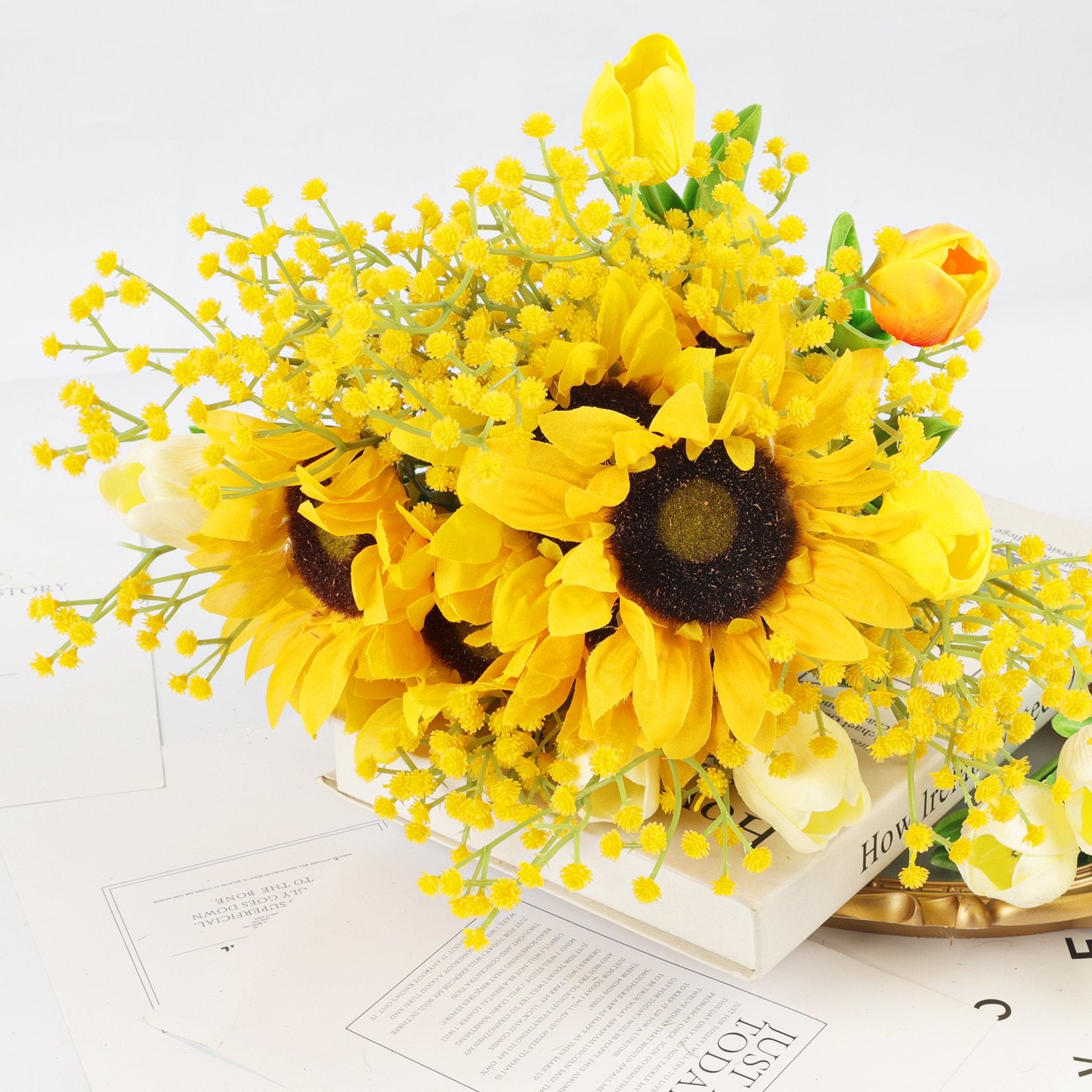 Artificial sunflower and tulip bouquet with yellow filler flowers arranged on books and papers.