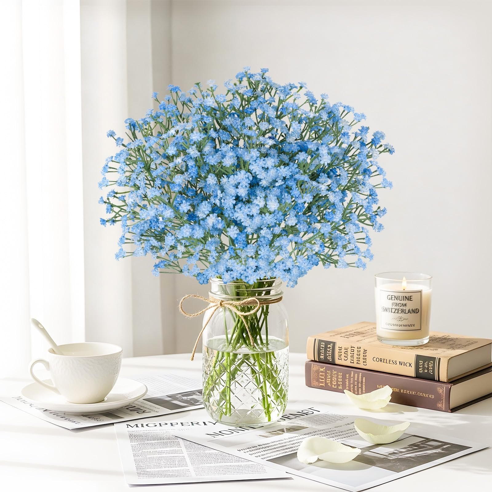Blue artificial baby's breath bouquet in a glass vase with a cup and books on a table