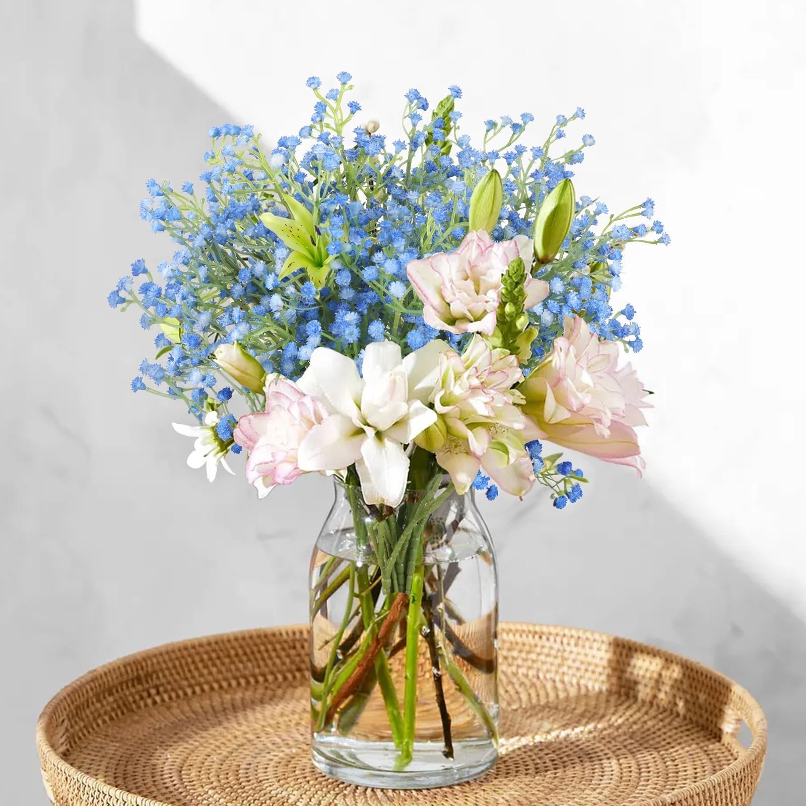 Bouquet of blue baby's breath and pink and white lilies in a glass vase on a woven tray.