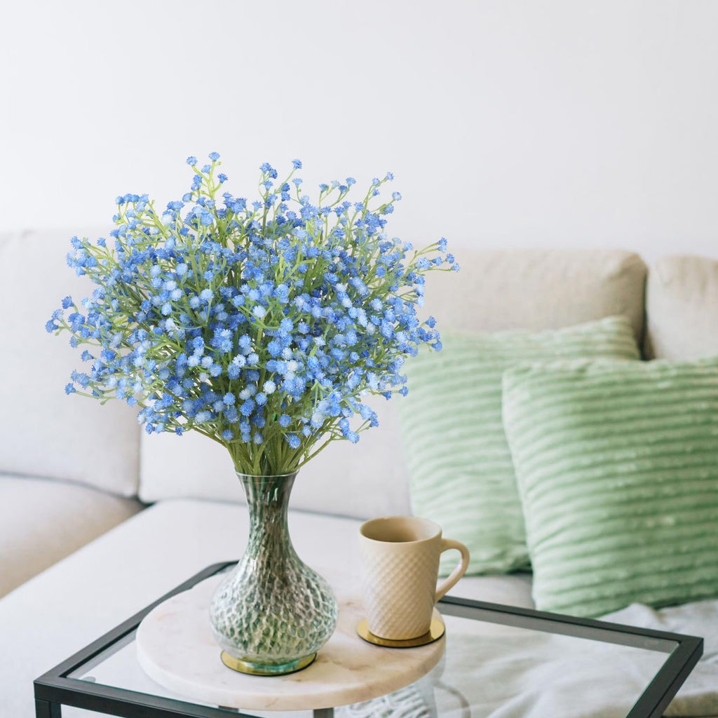Blue artificial baby's breath bouquet in a green vase on a table next to a mug, with a sofa and pillows in the background.