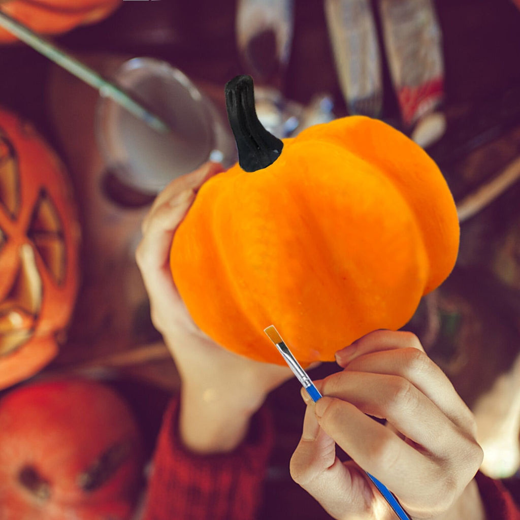 Hands painting an orange pumpkin with a brush, surrounded by other pumpkins and art supplies.