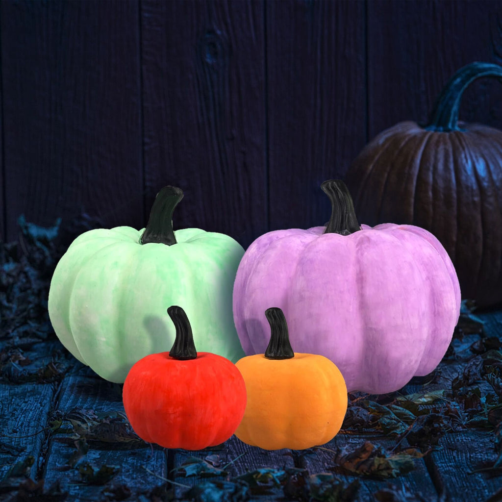 Four decorative pumpkins in various colors (green, purple, orange, red) arranged on a dark wooden surface with scattered leaves.