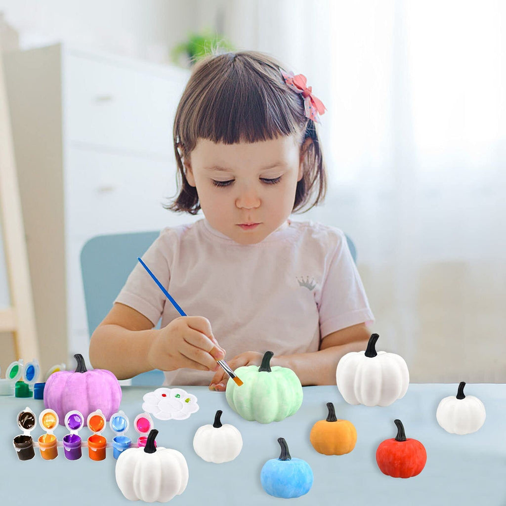 Young child painting a light green pumpkin with a brush, surrounded by other pumpkins and paints.