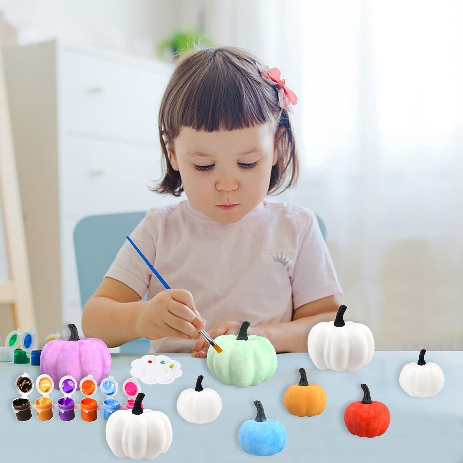 Young child painting a light green pumpkin with a brush, surrounded by other pumpkins and paints.