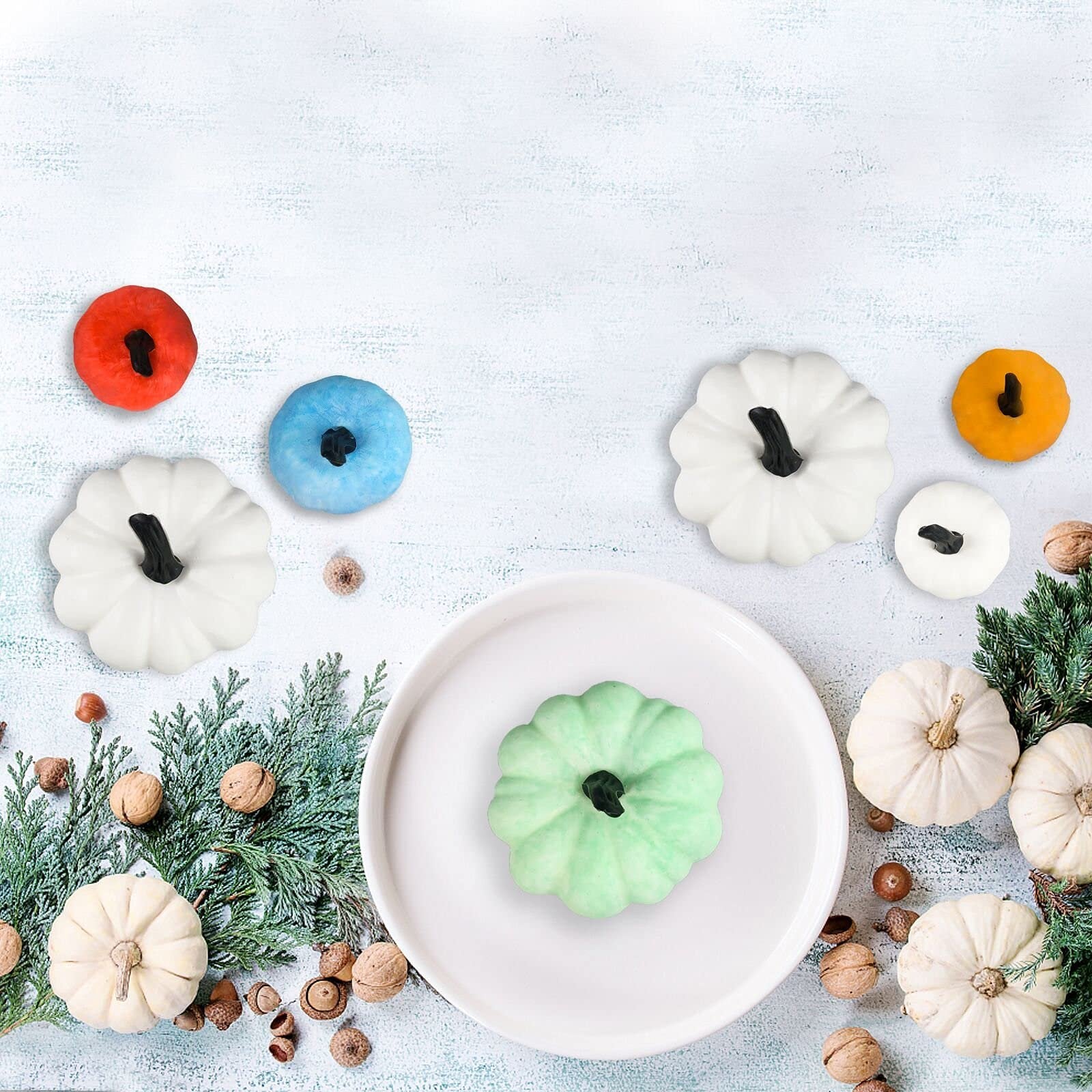 Assortment of decorative pumpkins in various colors and sizes arranged on a light blue textured surface with greenery and nuts.