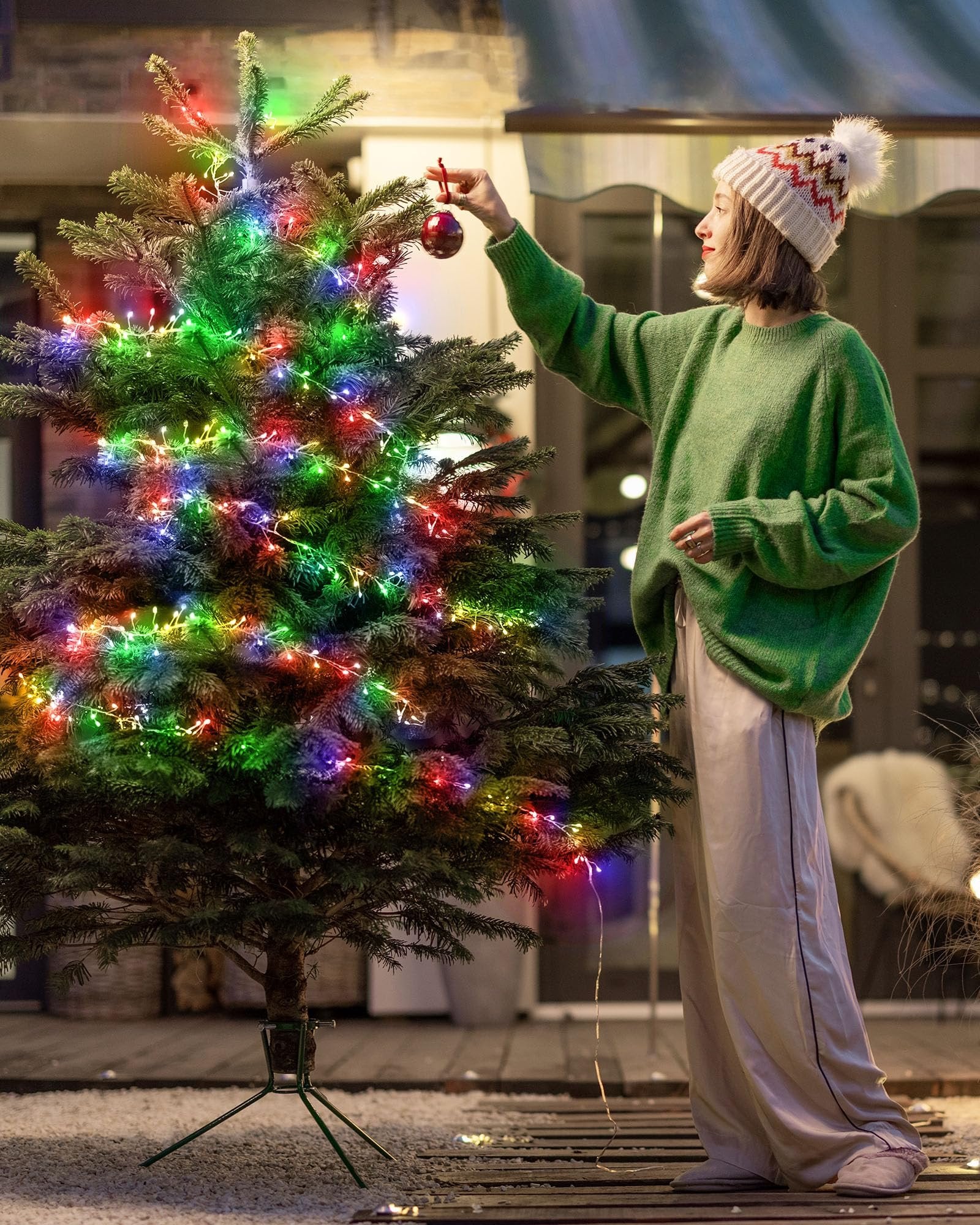 Woman in green sweater and patterned hat decorating a Christmas tree with multicolor fairy lights