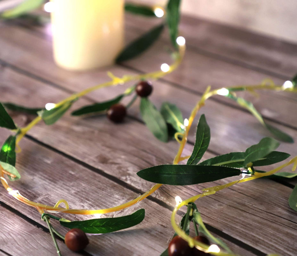 Olive branch garland string lights with warm white LEDs and faux olive fruits on a wooden surface with a blurred candle.
