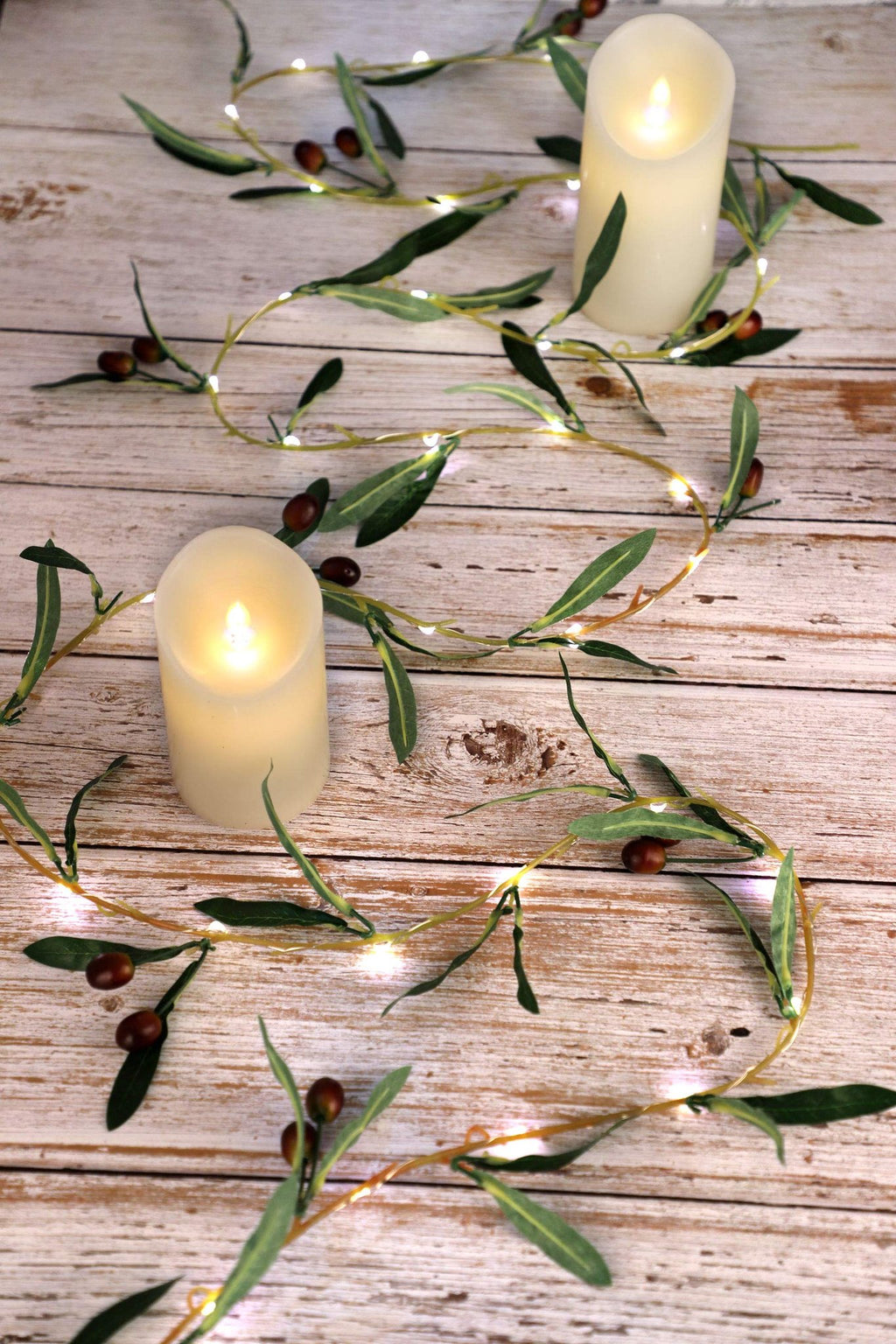 Olive branch garland string lights with warm white LEDs and faux olives draped around two pillar candles on a wooden surface.