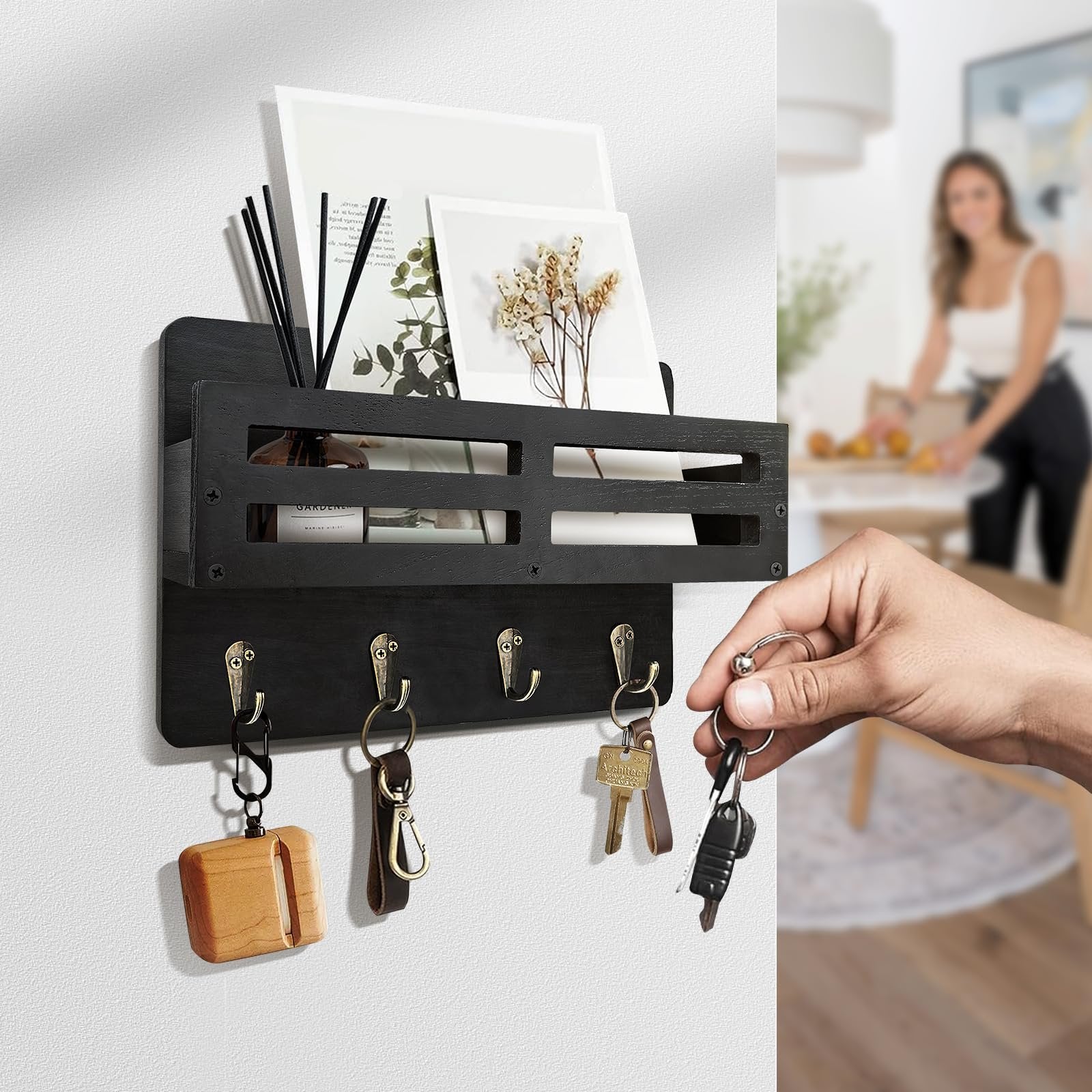 Black wooden key and mail holder with 4 hooks, a hand is adding keys to one hook. A woman is blurred in the background.