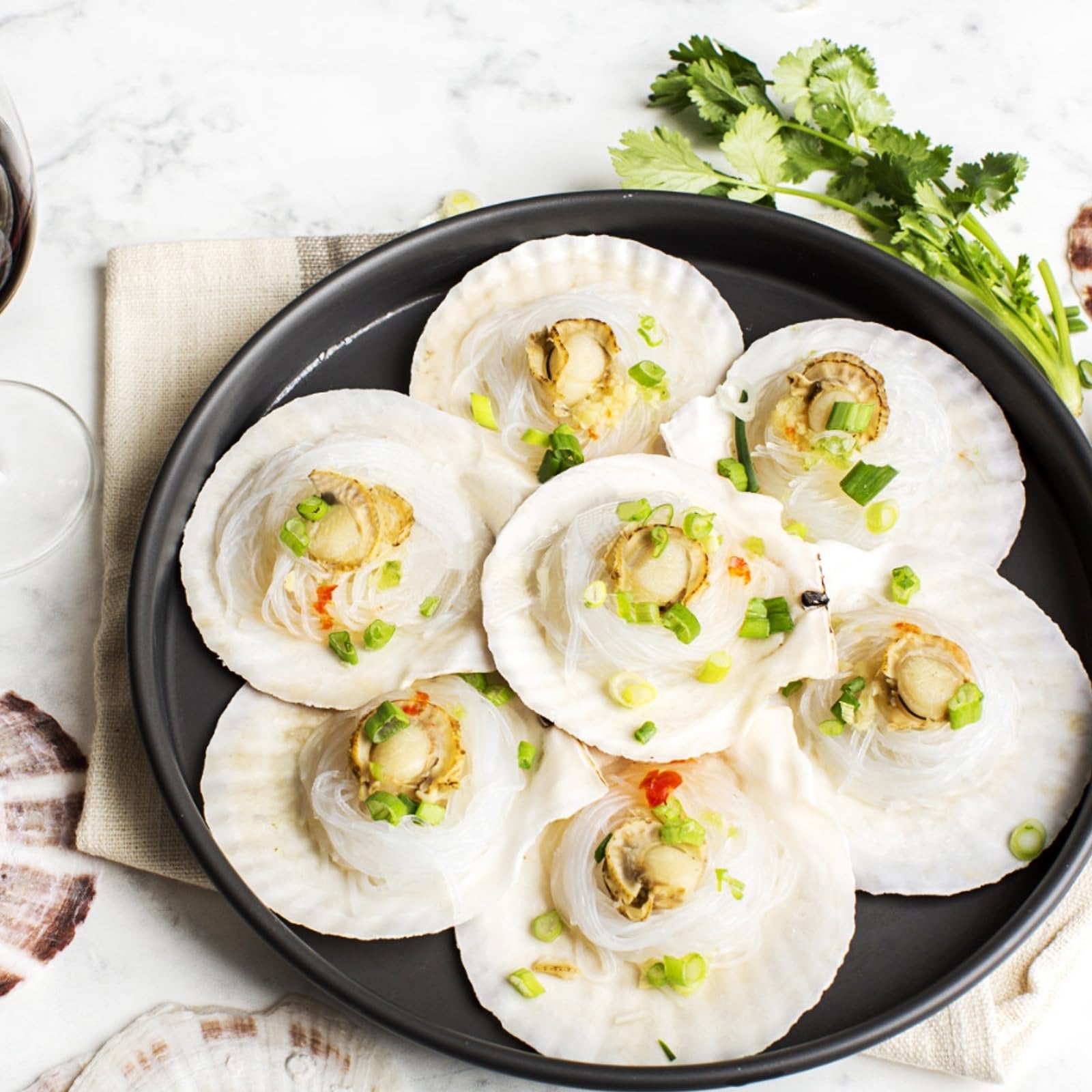 Steamed scallops in shells with noodles and green onions on a black plate, garnished with cilantro and a glass of red wine.
