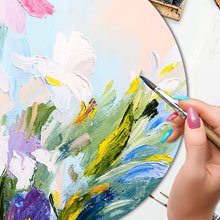 Artist's hand holding a paintbrush applying paint to a floral artwork on a wooden palette